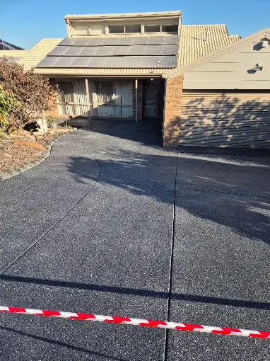 Freshly finished dark grey spray-on concrete driveway curving up to a brick carport, cordoned off with red and white barrier tape while it cures house with solar panels in the background. Spray-on concrete driveway by Spraycrete.