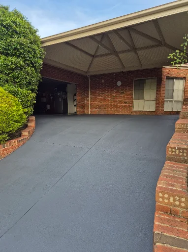 Wide grey spray-on concrete driveway with a smooth textured finish, curving toward a brick carport flanked by brick garden edging and lush greenery. Spray-on concrete driveway resurfacing by Spraycrete.